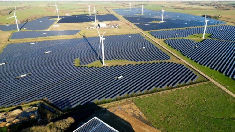 An aerial view of a large solar farm interspersed with wind turbines. Rows of solar panels cover vast areas of the land, while several tall wind turbines stand among them