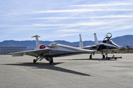 A NASA F-15 aircraft near X-59 with a white hangar and hills in the background, during electromagnetic interference testing.