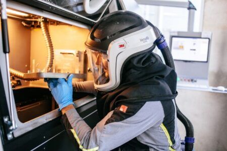 A technician wearing protective gear operates a metal additive manufacturing (3D printing) machine, handling a build plate with freshly printed components inside an industrial facility.