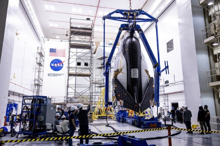 Dream Chaser spacecraft suspended vertically from lifting fixture inside NASA testing facility with technical personnel monitoring positioning operations