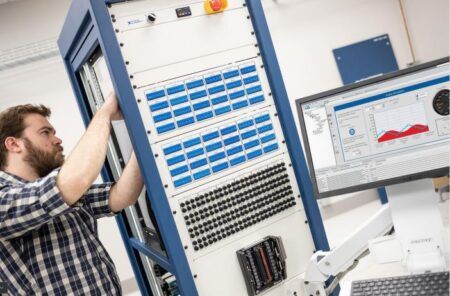 A technician in a plaid shirt adjusts an electronic test rack with multiple I/O modules, while a computer monitor displays a LabVIEW-based data analysis interface in a laboratory setting