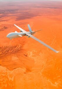 Alt text: White military drone flying over orange desert landscape with mountainous terrain