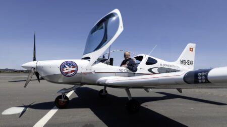 White two-seat electric aircraft with blue accents on airport tarmac with pilot visible in cockpit
