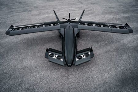 Gray military-style aircraft with tilting wing configuration and multiple propellers on tarmac