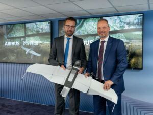 Two executives in business suits holding unmanned aircraft model in front of Airbus display screens