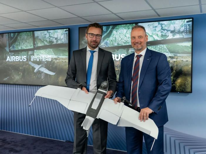 Two executives in business suits holding unmanned aircraft model in front of Airbus display screens