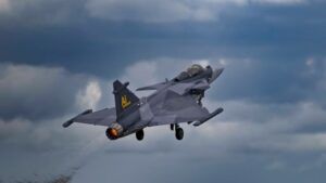 Gray Saab Gripen E fighter aircraft in flight against cloudy sky background with visible afterburner
