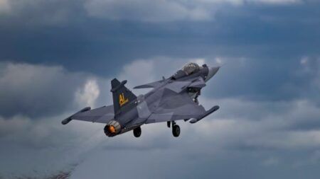Gray Saab Gripen E fighter aircraft in flight against cloudy sky background with visible afterburner