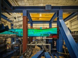 Large blue industrial testing frame with green monitoring display screens in aerospace structural testing laboratory