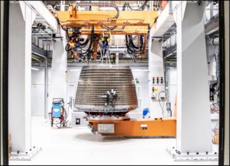 Large rocket engine component with metallic ribbed structure being manufactured in industrial facility with orange robotic equipment