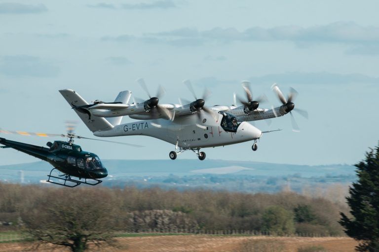 Vertical Aerospace eVTOL aircraft in flight during transition testing at Cotswold Airport with a chase helicopter visible in the background