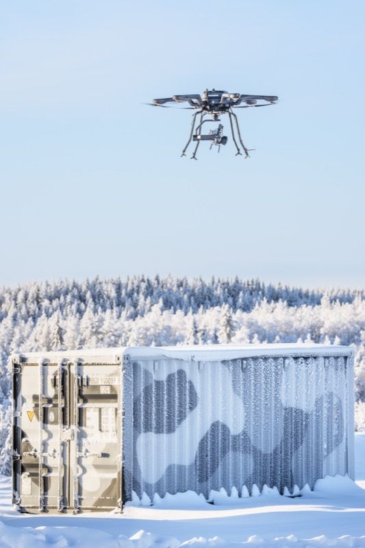 A multirotor drone hovering above a frost-covered shipping container at VTT's Arctic drone test site in northern Finland, with snow-laden forest in the background