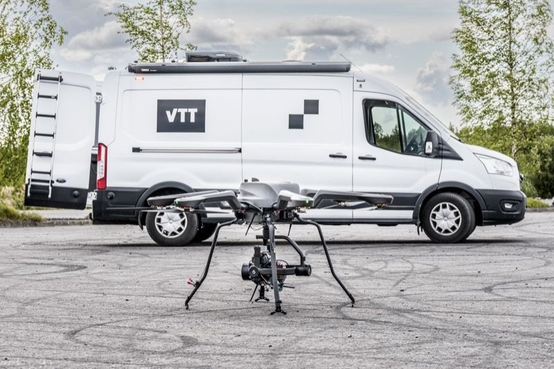 A multirotor drone positioned on the ground in front of a VTT-branded mobile operations vehicle at the Finnish test site