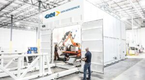 Large-scale additive manufacturing cell at GKN Aerospace's Fort Worth facility, with a robotic arm visible inside the enclosure and two technicians operating equipment