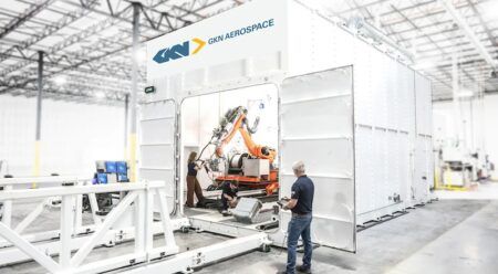 Large-scale additive manufacturing cell at GKN Aerospace's Fort Worth facility, with a robotic arm visible inside the enclosure and two technicians operating equipment