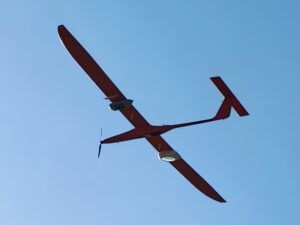 Underside view of a dark red fixed-wing uncrewed aircraft in flight against a clear blue sky, with two cylindrical pod-mounted payloads carried beneath the wings