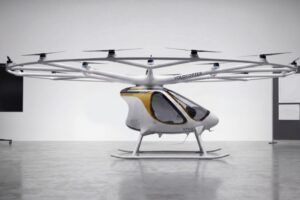 Volocopter VoloXPro two-seat ultralight electric multicopter in a studio setting, showing its ring of 18 rotors on radial arms above a white and gold enclosed cabin with skid undercarriage