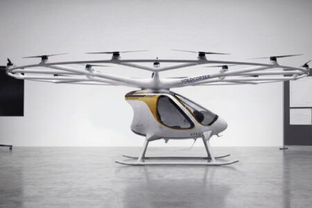 Volocopter VoloXPro two-seat ultralight electric multicopter in a studio setting, showing its ring of 18 rotors on radial arms above a white and gold enclosed cabin with skid undercarriage