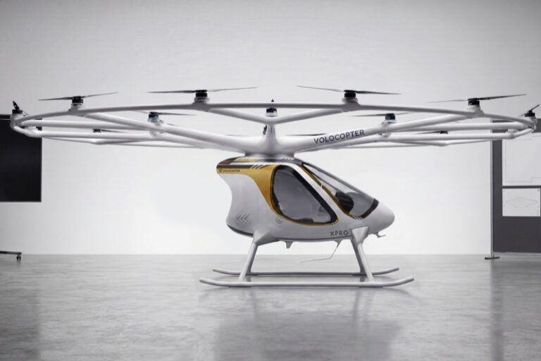 Volocopter VoloXPro two-seat ultralight electric multicopter in a studio setting, showing its ring of 18 rotors on radial arms above a white and gold enclosed cabin with skid undercarriage