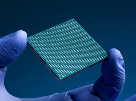 NASA tests more powerful spacecraft processor to enable onboard AI A gloved hand holds a teal square semiconductor chip showing a dense grid of silver solder bumps on its underside against a dark blue background