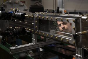 A researcher looks through the viewing window of a supersonic wind tunnel test section, with high-speed cameras and optical diagnostic equipment mounted alongside the metal-bolted chamber
