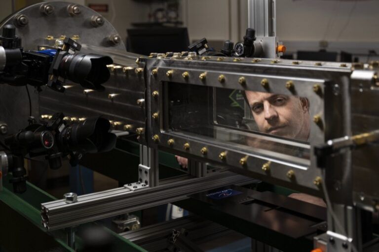 A researcher looks through the viewing window of a supersonic wind tunnel test section, with high-speed cameras and optical diagnostic equipment mounted alongside the metal-bolted chamber