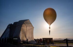 A large balloon ascends at sunrise carrying a cone-shaped flight test payload above a desert launch site