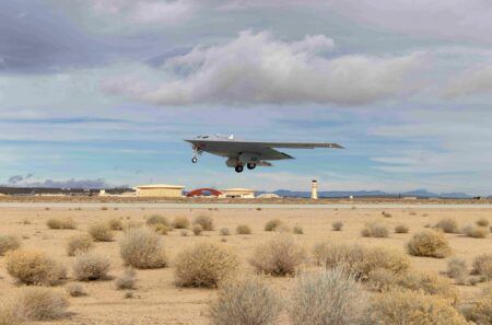 US Air Force combines operational and developmental flight testing on B-21 Raider program Alt text: A B-21 Raider stealth bomber in low-level flight above an arid desert landscape at Edwards Air Force Base, with hangars and a control tower visible in the background