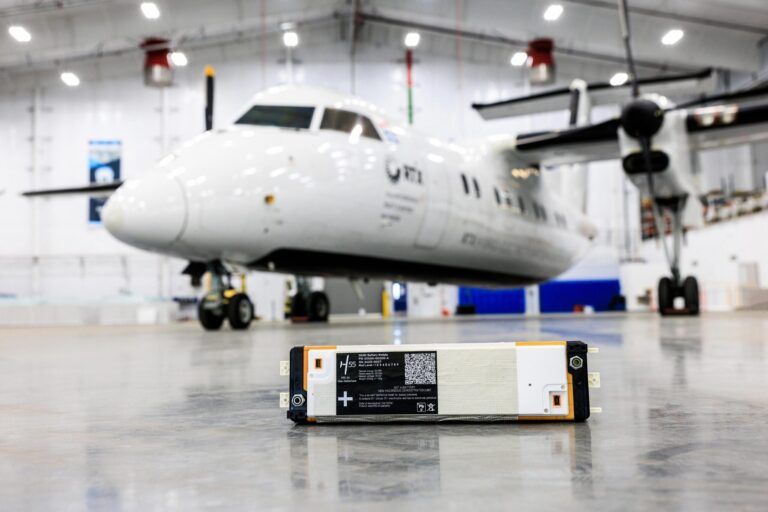An H55 Adagio battery module on a hangar floor, with the RTX Hybrid-Electric Flight Demonstrator turboprop aircraft visible in the background