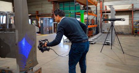 Technician using a handheld 3D scanner emitting blue structured light on a large metal aerospace component in an industrial workshop, with a laser tracker on a tripod in the background