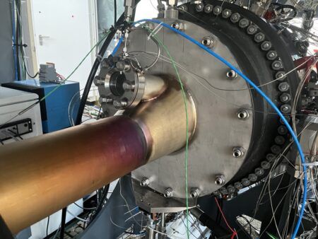 Hydrogen sector combustor test rig at DLR Institute of Space Propulsion, Lampoldshausen, showing heat-discolored fuel inlet pipe connected to a circular test chamber flanked by instrumentation cabling