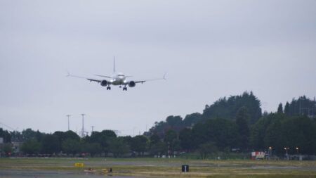 A Boeing 737-10 test aircraft on final approach to land at Boeing Field on an overcast day, with landing gear extended over the airfield perimeter
