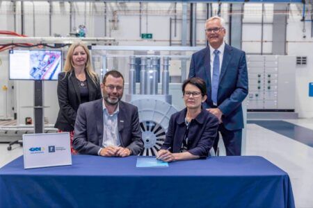 GKN Aerospace launches startup to develop cryogenic motors for aircraft Two people sign documents at a blue-draped table bearing GKN Aerospace and University of Nottingham logos, with two colleagues standing behind and a large electrical machine test rig in a transparent enclosure in the background