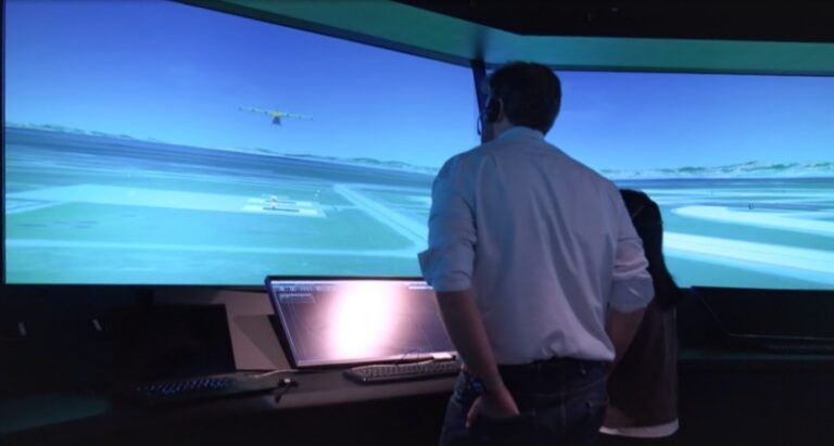Wisk and NASA test one supervisor for three autonomous aircraft Two people standing at a workstation watching a wide curved screen that displays a simulated airport, with an autonomous aircraft in flight and another on the runway