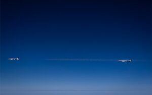Two business jets flying in close formation at high altitude, with a thin contrail trailing between them against a deep blue sky