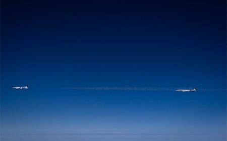 Two business jets flying in close formation at high altitude, with a thin contrail trailing between them against a deep blue sky