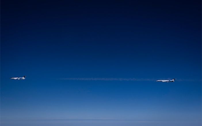Two business jets flying in close formation at high altitude, with a thin contrail trailing between them against a deep blue sky