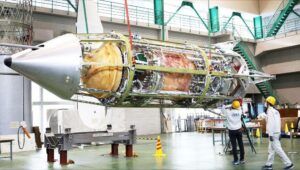 The cylindrical RV-X test vehicle lying horizontally in an assembly hall with its outer panels removed to expose internal propellant tanks, plumbing and the engine bay, a conical nose at left, as two technicians in hard hats work alongside