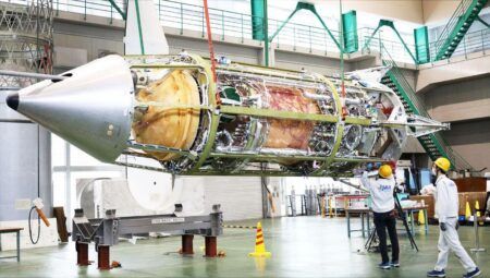 The cylindrical RV-X test vehicle lying horizontally in an assembly hall with its outer panels removed to expose internal propellant tanks, plumbing and the engine bay, a conical nose at left, as two technicians in hard hats work alongside