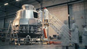 Engineers in cleanroom suits working on a metallic spacecraft capsule mounted on a test stand in a manufacturing facility