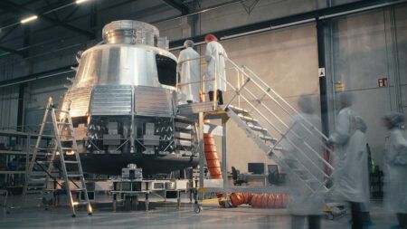 Engineers in cleanroom suits working on a metallic spacecraft capsule mounted on a test stand in a manufacturing facility