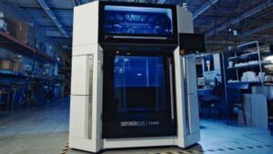 Stratasys F3300 industrial 3D printer with its build chamber illuminated in blue light on a factory floor