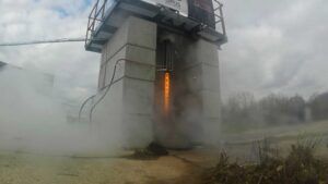 A rocket engine test stand with an orange flame and thick exhaust smoke rising from a concrete gantry