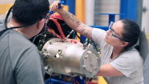 Two technicians in safety glasses fitting instrumentation and wiring to a cylindrical metal power and cooling unit in a test facility