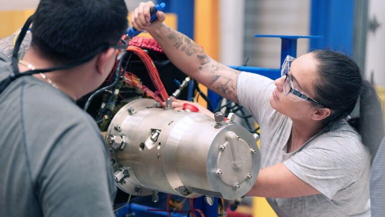 Two technicians in safety glasses fitting instrumentation and wiring to a cylindrical metal power and cooling unit in a test facility
