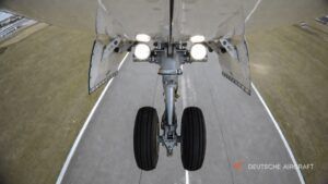 View from beneath a D328 aircraft looking at its extended twin-wheel nose landing gear, with the gear-bay doors open and landing lights illuminated above the runway