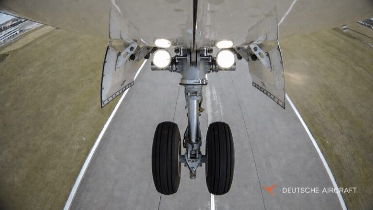 View from beneath a D328 aircraft looking at its extended twin-wheel nose landing gear, with the gear-bay doors open and landing lights illuminated above the runway