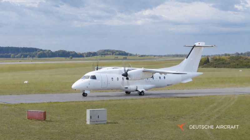 A white D328 twin-turboprop regional aircraft on a taxiway at an airfield, with grass and trees in the background