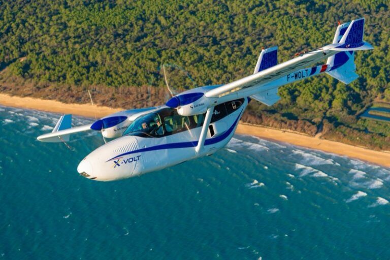 The X-VOLT hybrid-electric demonstrator in flight over a coastline, showing its twin-boom airframe and rear-mounted propulsion