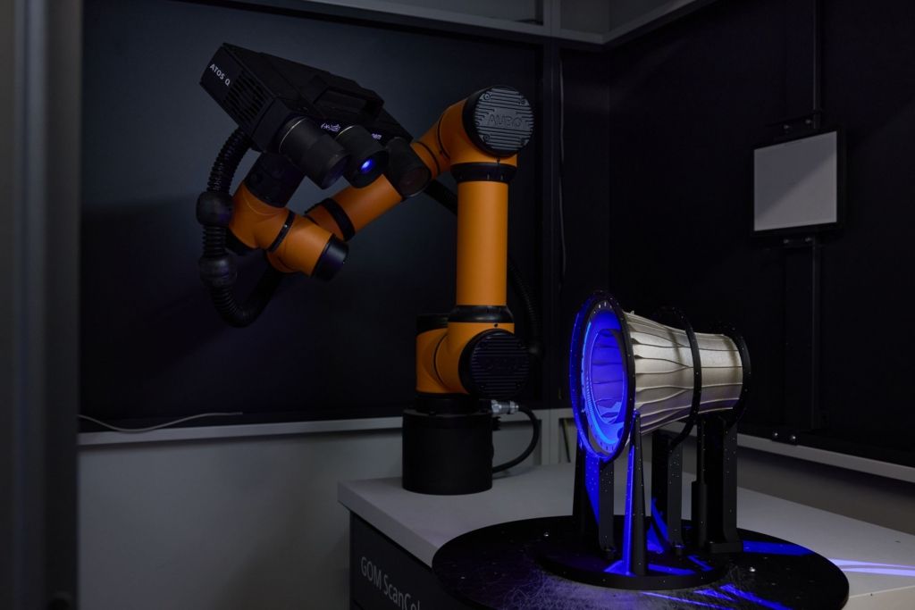 Orange robotic arm fitted with a structured-light scanner inspecting a metallic additively manufactured engine component lit in blue